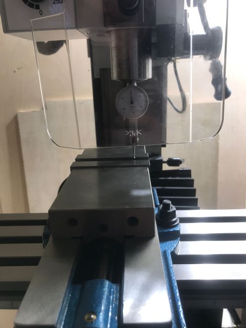 Image of a milling machine from the front. There is a dial test indicator mounted in the spindle via a 3/8" chuck; the indicator's face is showing. Its tip is on the top edge of the fixed jaw of a blue-and-unfinished-steel milling vise mounted on the milling table.