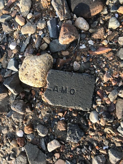 A cobble made of a cinder-block-ish material, about the dimensions of a half-brick, with one broken side and three weathered square sides. On the face it has the end of some text reading "LAMO".