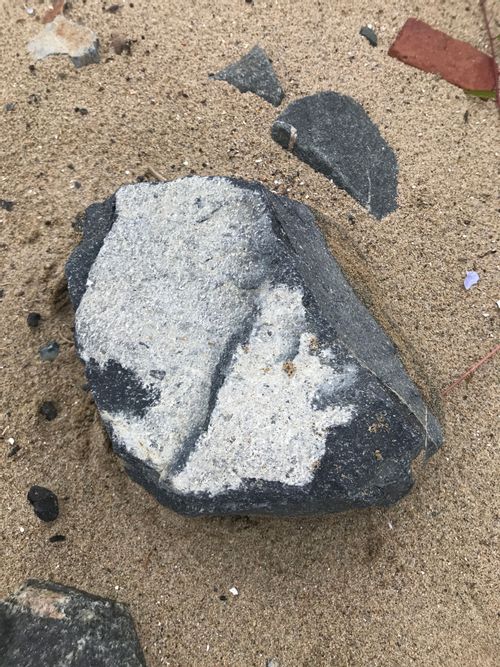 A moderately-large cobble of the dark-grey material, broadly cube-shaped. A brick in the background suggests that it's around 12" x 10" X 8". It has a white cortex on the side facing the viewer. 