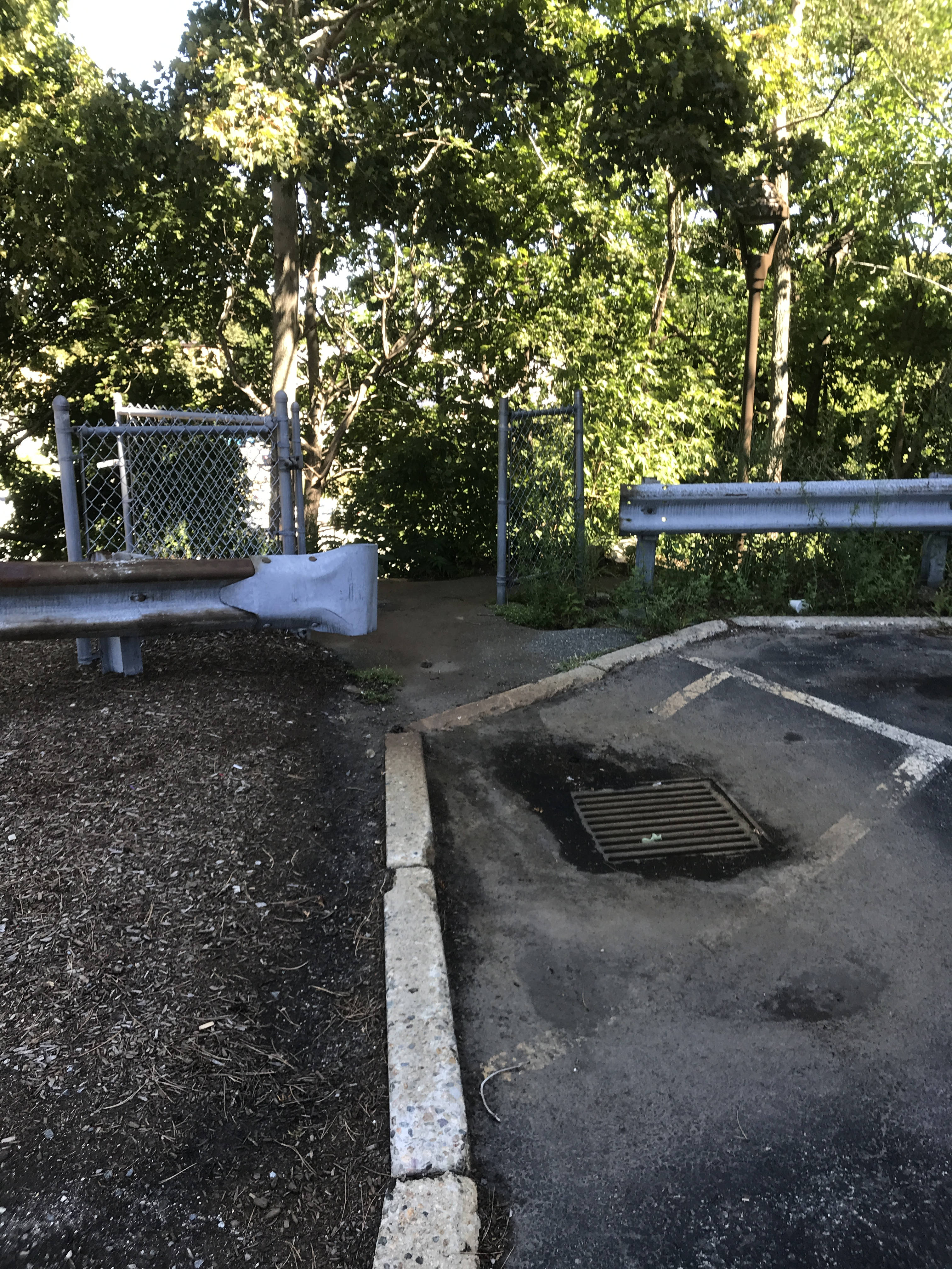 A small break between two guard rails reveals two freestanding sections of fence with an open gate between them. In front of the opening is a storm drain with a muddy puddle drying around it.