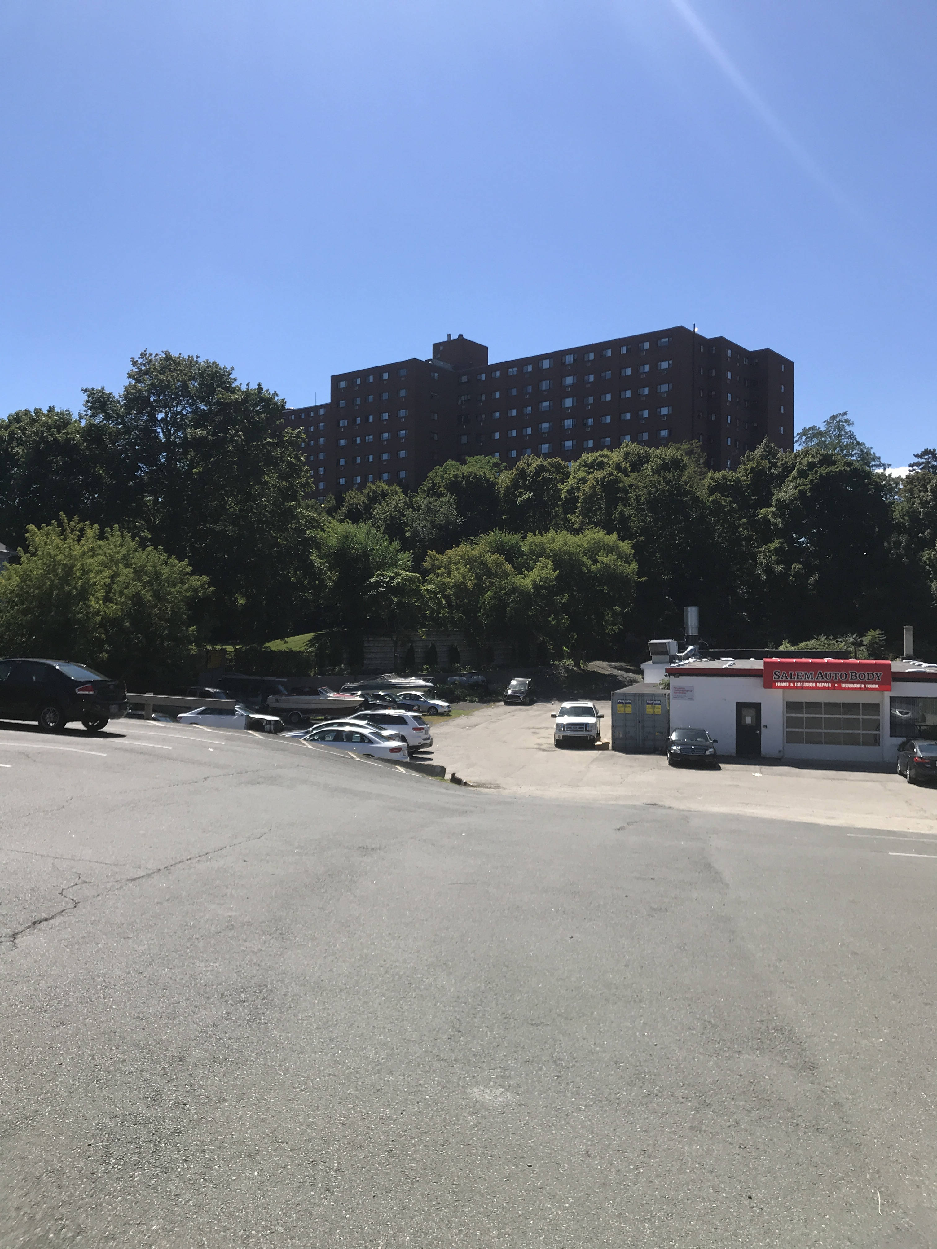 A view across a street. In the middle ground is an auto-body shop surrounded by a sizeable lot with a few cars parked in it. Behind the lot is a wooded area, and behind that, six stories of a long dark brick building rise over the trees.