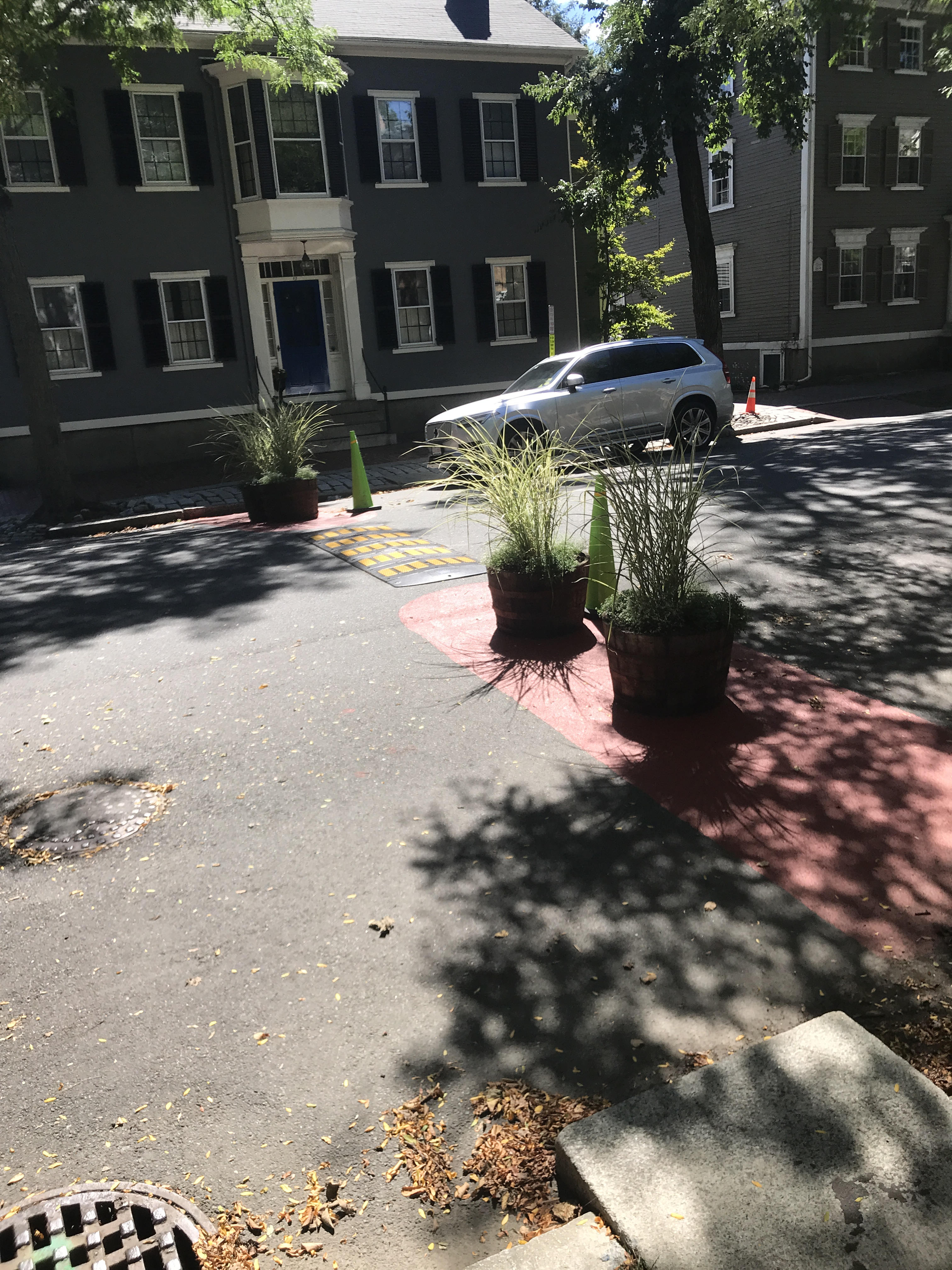 Across a wide road, ornamental planters and green caution cones leave only a single car-width passable to cars as a traffic-calming measure. A non-permanent speed bump is stretched over the opening