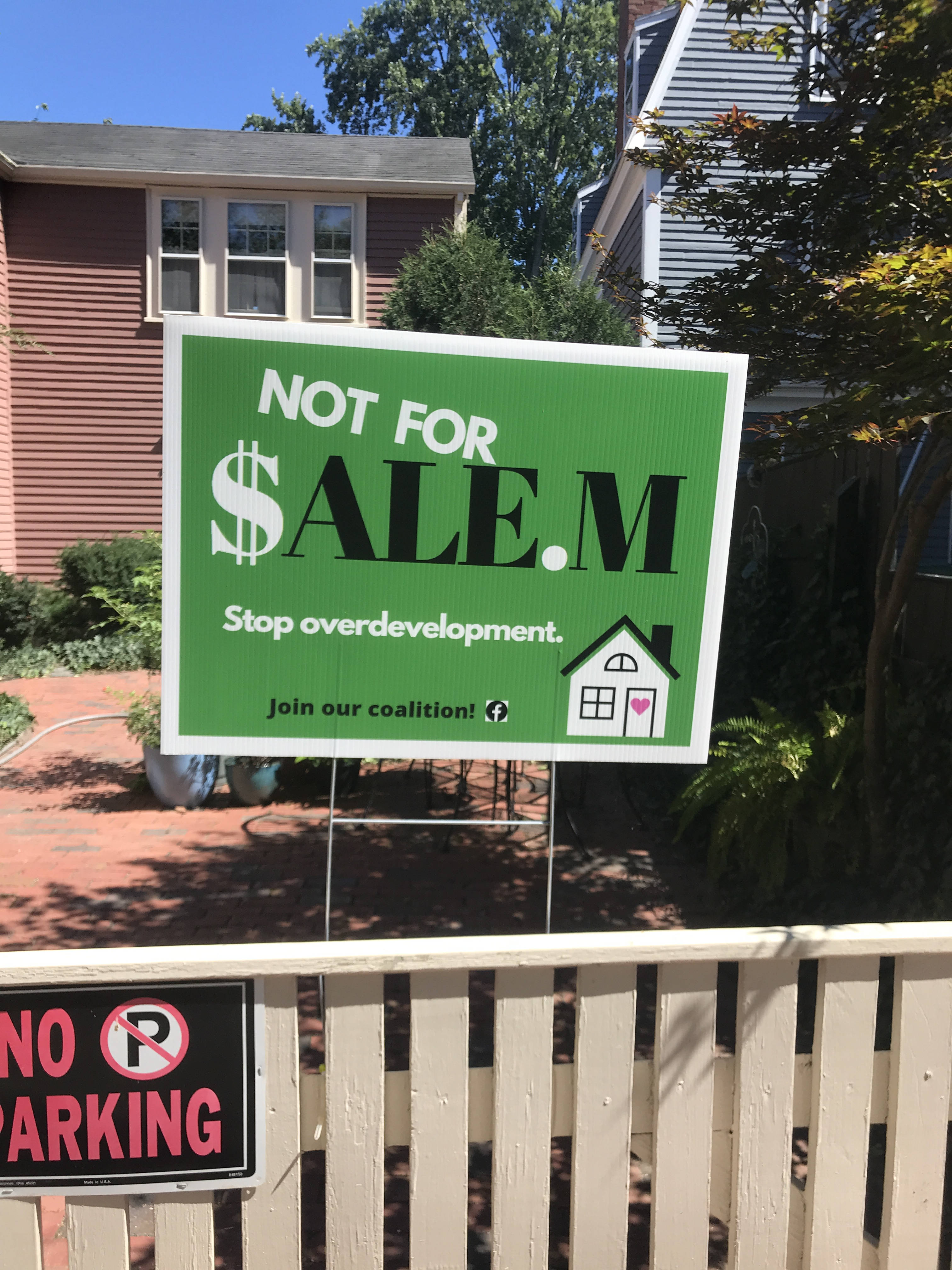 A cream-colored fence in front of an antique home. The fence bears a 'No Parking' sign and one of the 'Not For $alem' signs. The 'Not for $alem' sign says 'Stop Overdevelopment. Join our coalition!' and has a picture of a single-family house on it. The door of the house is decorated with a heart.