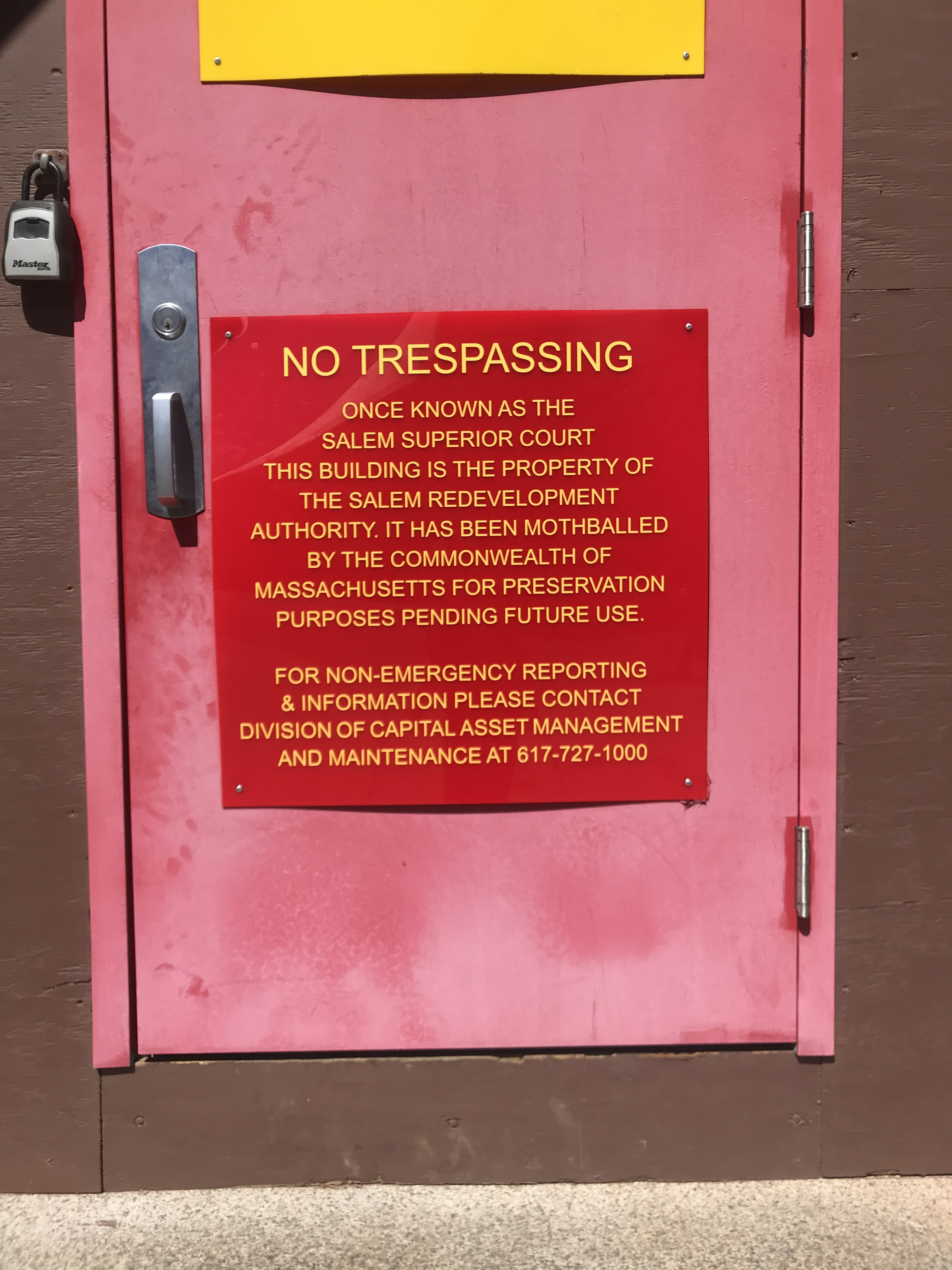 A shiny plastic sign on a faded red-painted door reads 'NO TRESPASSING / Once known as the Salem Superior Court / this building is the property of the Salem Redevelopment Authority. It has been mothballed by the Commonwealth of Massachusetts for preservation purposes pending future use/ For non-emergency reporting & information please contact division of capital asset management and maintenance at 617-727-1000'