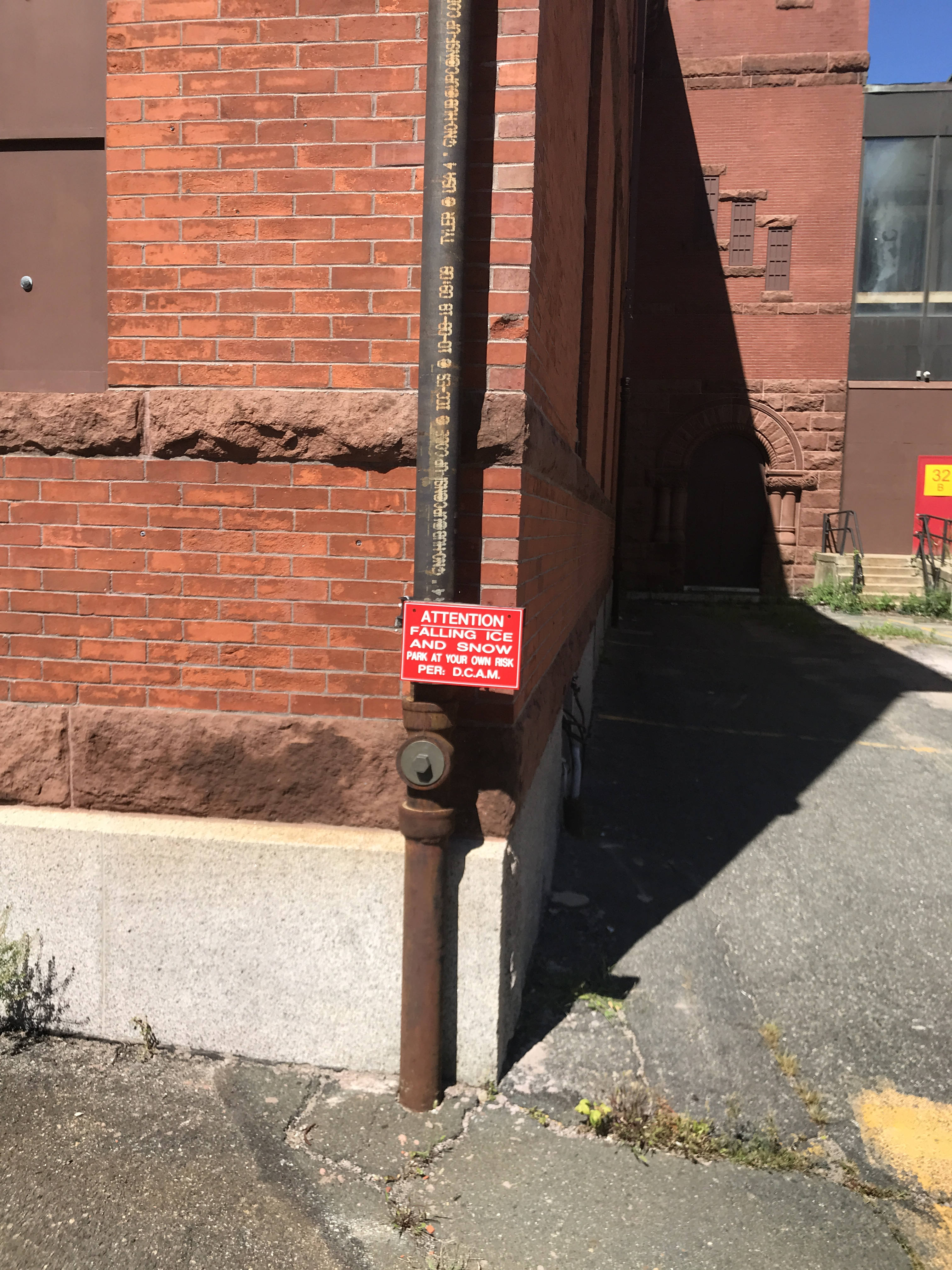 at a corner of a brick building, a bright red sign attached to a drainpipe reads 'ATTENTION: Falling Ice And Snow, Park At Your Own Risk. Per D.C.A.M